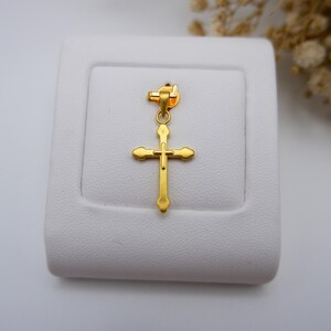 999 pure gold cross pendant displayed on a white leatherette jewelry stand, emphasizing its gleaming yellow tone and minimalist hollow design. The lightweight crucifix charm features soft, rounded ends and a polished finish, making it suitable for both men and women. Dried white flowers add a soft, neutral backdrop, enhancing the elegance. Ideal for search terms like real gold cross necklace charm, Catholic pendant, gold faith jewelry, or spiritual gift idea.