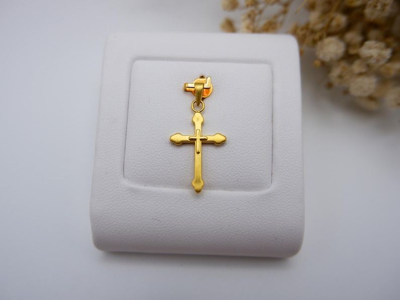 999 pure gold cross pendant displayed on a white leatherette jewelry stand, emphasizing its gleaming yellow tone and minimalist hollow design. The lightweight crucifix charm features soft, rounded ends and a polished finish, making it suitable for both men and women. Dried white flowers add a soft, neutral backdrop, enhancing the elegance. Ideal for search terms like real gold cross necklace charm, Catholic pendant, gold faith jewelry, or spiritual gift idea.