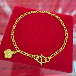 May include: A gold chain bracelet featuring a series of elongated oval links and a single five-petaled flower charm. The bracelet is secured with an S-hook clasp, and the metal has a bright, polished yellow gold finish throughout the entire piece.