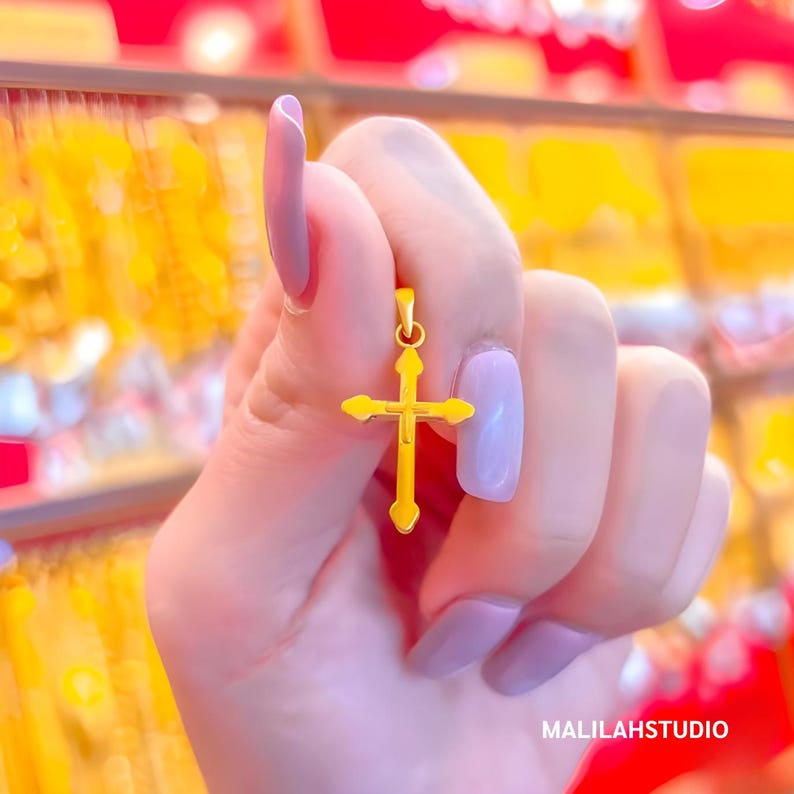 Close-up of a woman's hand holding a minimalist gold cross pendant in a vibrant jewelry store. The pendant is crafted from 999 pure gold with heart-shaped tips and a polished finish, offering lightweight elegance. The pastel manicure contrasts with the red-yellow background, making the gold pop. Perfect for Christian gifts, spiritual jewelry, or minimalist style. Branded with MALILAH STUDIO for a premium touch.
