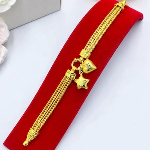 May include: A gold bracelet featuring a double-strand woven chain design. The bracelet includes two dangling charms, one shaped like a heart and the other shaped like a five-pointed star, both with a textured finish and a bright yellow gold tone.