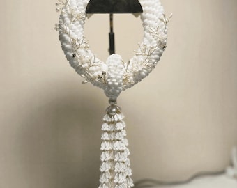 Könnte beinhalten: Ein kreisförmiger weißer Blumenschmuck aus dicht angeordneten kleinen weißen Blütenblättern, akzentuiert durch zarte Drahtzweige mit winzigen weißen Perlen. Eine lange, gestufte weiße Quaste hängt an der Unterseite herab.