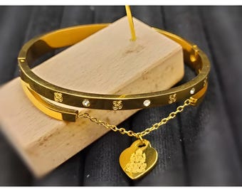 Könnte beinhalten: Ein goldenes Armreif-Armband mit einem herzförmigen Anhänger. Das Armband ist mit kleinen, klaren Steinen und dem Om-Symbol verziert. Der Anhänger ist mit einer detaillierten Gravur einer Gottheit versehen.