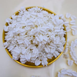 A large collection of cream white crown flower beads for bulk lei supply, displayed in a golden tray on a white fabric background.