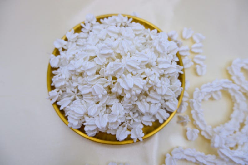 A large collection of cream white crown flower beads for bulk lei supply, displayed in a golden tray on a white fabric background.