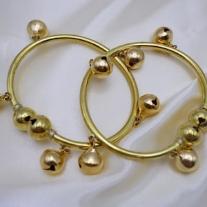 May include: Two gold-colored bangles with attached bells. Each bangle has a circular design with multiple small bells and larger bell-shaped charms. The bangles are displayed on a white, silky fabric.