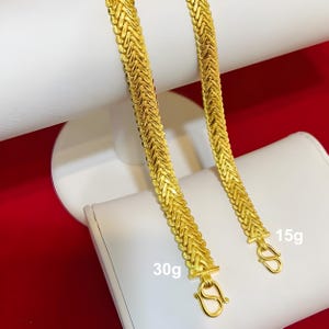 K&ouml;nnte beinhalten: Zwei goldene Ketten, eine mit 15 Gramm und die andere mit 30 Gramm, werden auf einer wei&szlig;en Oberfl&auml;che pr&auml;sentiert. Die Ketten sind aus einem gewebten Muster gefertigt und haben einen Verschluss am Ende.