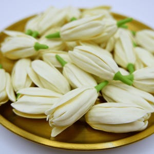 May include: A collection of ivory-colored flower buds with green stems, arranged on a gold-colored plate. The buds are tightly closed, suggesting they are about to bloom. The plate has a simple, round design.