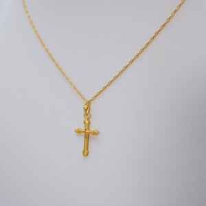 May include: A gold-colored necklace with a cross pendant. The cross is a simple design with rounded edges. The chain is a delicate, twisted rope style. The necklace is displayed against a white background.
