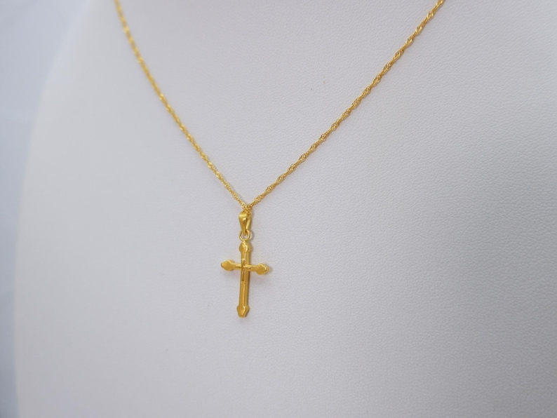 May include: A gold-colored necklace with a cross pendant. The cross is a simple design with rounded edges. The chain is a delicate, twisted rope style. The necklace is displayed against a white background.