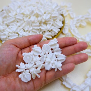 Close-up of a hand holding several white faux crown flower beads to show scale and realistic floral texture for DIY crafts.