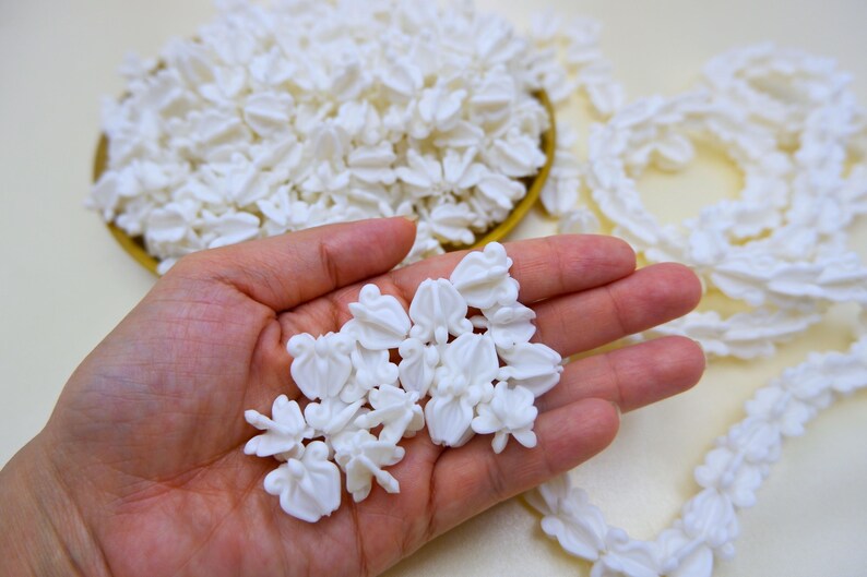 Close-up of a hand holding several white faux crown flower beads to show scale and realistic floral texture for DIY crafts.