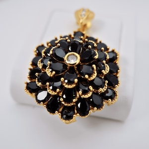 May include: A gold-toned pendant featuring a floral design. The pendant is adorned with numerous black, teardrop-shaped gemstones, arranged in layers. A single, clear gemstone is centered, and the bail is embellished with small clear stones.
