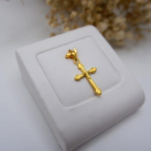 Close-up of a 999 pure gold cross pendant resting on a white leather jewelry stand, showcasing its elegant hollow design and polished yellow gold surface. The crucifix charm features rounded heart-like edges and a delicate bail, ideal for fine necklaces. Dried flowers in the background add a soft, natural contrast, making this image perfect for SEO terms like real gold Catholic pendant, Christian cross charm, dainty religious necklace, or spiritual gold gift.