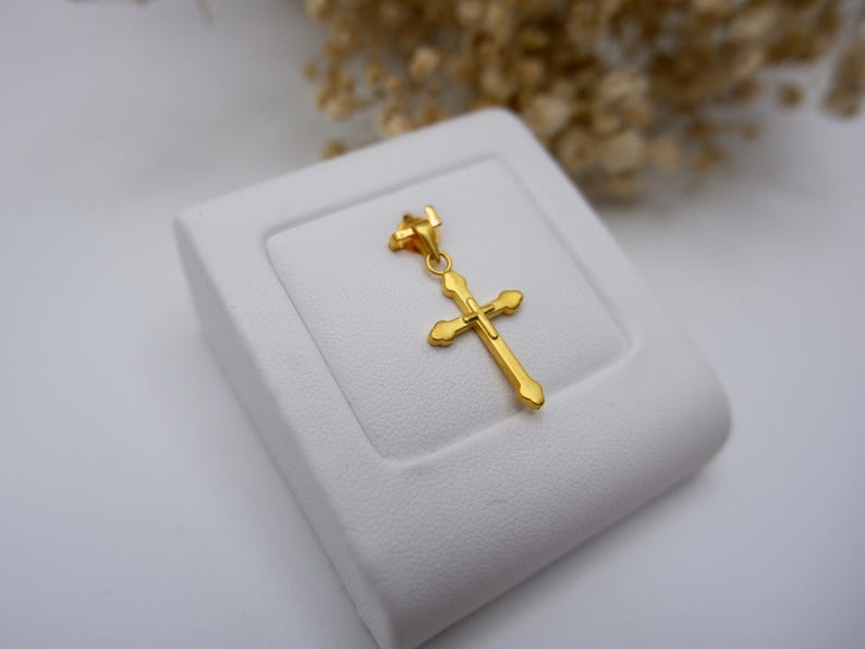 Close-up of a 999 pure gold cross pendant resting on a white leather jewelry stand, showcasing its elegant hollow design and polished yellow gold surface. The crucifix charm features rounded heart-like edges and a delicate bail, ideal for fine necklaces. Dried flowers in the background add a soft, natural contrast, making this image perfect for SEO terms like real gold Catholic pendant, Christian cross charm, dainty religious necklace, or spiritual gold gift.