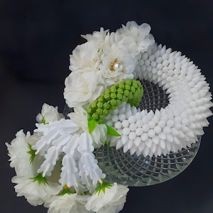 May include: A white floral garland with a green accent and a pearl-centered embellishment. The garland is arranged on a clear, patterned glass dish against a dark background. The flowers are clustered and layered.