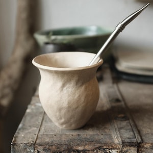 Könnte beinhalten: Eine hellbraune Keramik-Mate-Kürbis mit einer strukturierten Oberfläche. Der Kürbis steht auf einer rustikalen Holzoberfläche mit einem Metall-Bombilla-Strohhalm.