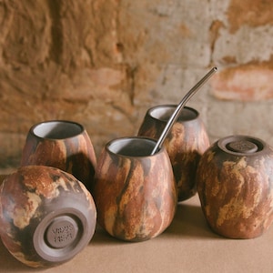 Puede incluir: Colección de cinco calabazas de yerba mate hechas a mano con una estética natural y terrosa. Las calabazas presentan un patrón veteado marrón y beige, con una pajita de acero inoxidable en una de ellas. La base de una calabaza tiene el texto "VIDUS REA".