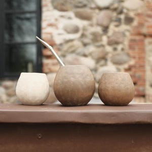 以下が含まれることがあります： 3つのひょうたん型の容器があり、1つは薄いベージュ、2つは茶色で、茶色の表面に置かれています。金属製のストローが中央の容器に入っています。背景には石壁と暗い窓があります。