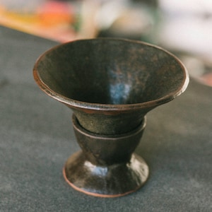 Può includere: Un vaso in ceramica verde scuro fatto a mano con una parte superiore ampia e svasata e una base arrotondata. La ceramica ha una superficie strutturata e un bordo marrone rossiccio. L'altezza totale è di circa 13 cm.