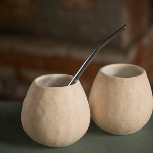 Può includere: Due tazze in ceramica beige con una superficie testurizzata e una cannuccia in metallo argentato.