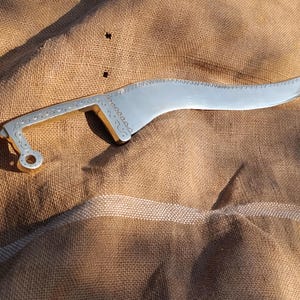 May include: A silver-colored, curved blade with a decorative handle. The blade has a serrated edge and a pointed tip. The handle features intricate etched designs and a circular opening at the end. The object is set against a brown, textured background.