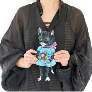 Puede incluir: Un reloj de pared con forma de gato negro que lleva un vestido de cuadros azul claro con un lazo rosa. El gato sostiene una pequeña muñeca en sus brazos. La esfera del reloj está integrada en el vestido, con manecillas negras y números.