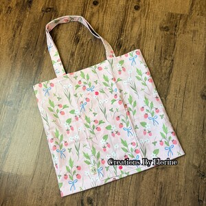 以下が含まれることがあります： イチゴと花の模様が入ったピンク色のトートバッグ。バッグにはボタン留めとハンドルが付いています。デザインにはイチゴ、緑の葉、白い花、青いリボンが含まれています。「Creations By Florine」の文字がバッグに表示されています。
