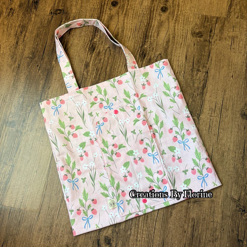 以下が含まれることがあります： イチゴと花の模様が入ったピンク色のトートバッグ。バッグにはボタン留めとハンドルが付いています。デザインにはイチゴ、緑の葉、白い花、青いリボンが含まれています。「Creations By Florine」の文字がバッグに表示されています。