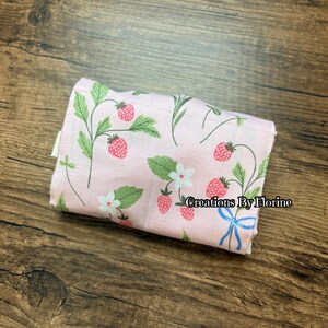 以下が含まれることがあります： イチゴ柄のピンク色の折り畳まれた布製品。赤いイチゴ、緑の葉、白い花が特徴です。「Creations By Florine」の文字が見えます。布は木製の表面にあります。