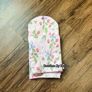 以下が含まれることがあります： イチゴと花の模様のピンク色の布製品で、緑の葉、赤いイチゴ、青いリボンが特徴です。商品は折りたたまれ、ピンク色のボタンで留められています。「Creations By Florine」の文字が見えます。