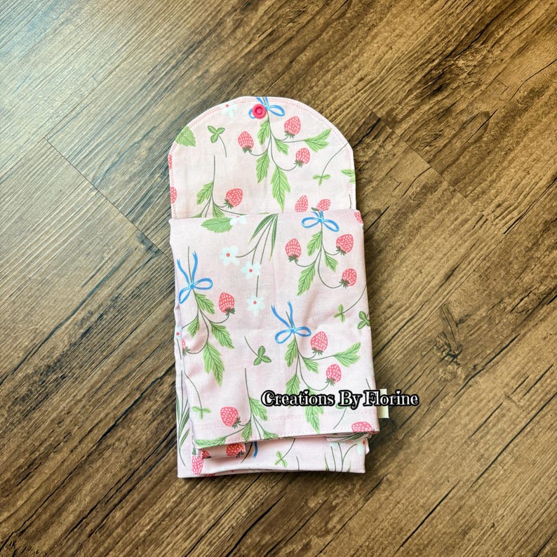 以下が含まれることがあります： イチゴと花の模様のピンク色の布製品で、緑の葉、赤いイチゴ、青いリボンが特徴です。商品は折りたたまれ、ピンク色のボタンで留められています。「Creations By Florine」の文字が見えます。