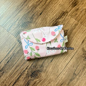 以下が含まれることがあります： イチゴと花の模様、青いリボンがアクセントになった、フラップ開閉式のピンク色の布製財布。財布はピンク色のボタンで留められています。「Creations By Florine」の文字が財布にプリントされています。