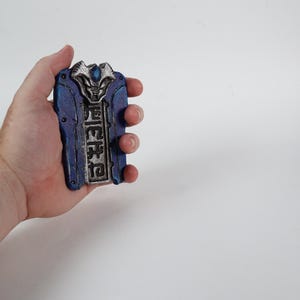 以下が含まれることがあります： 手で持たれた、青と銀の装飾品。精巧なディテールが施されており、長方形の形状に様式化されたデザインとテキストが施されています。配色は青、銀、その他の色のヒントを含んでいます。