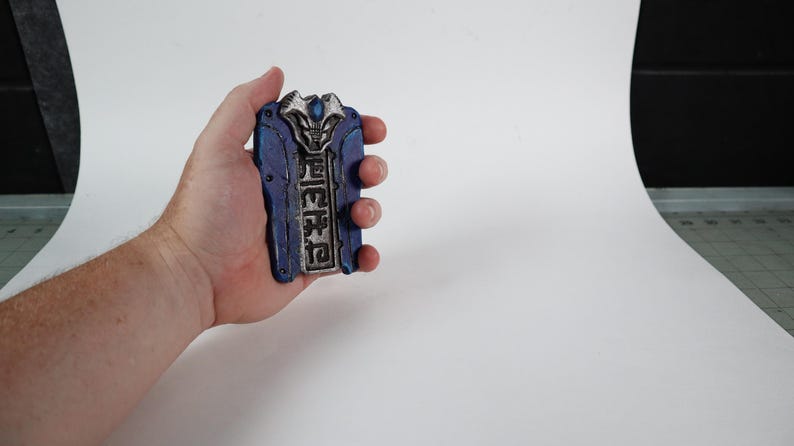 以下が含まれることがあります： 手で持たれた、青と銀の装飾品。精巧なディテールが施されており、長方形の形状に様式化されたデザインとテキストが施されています。配色は青、銀、その他の色のヒントを含んでいます。