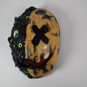 Op de afbeelding: Een tweekleurig masker met een griezelig ontwerp. De linkerkant is zwart met meerdere groene ogen. De rechterkant is bruin met een zwarte kruis en een rode mesh mond. Het masker heeft een verweerde, getextureerde uitstraling.