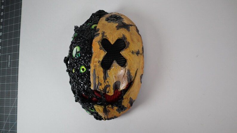 Op de afbeelding: Een tweekleurig masker met een griezelig ontwerp. De linkerkant is zwart met meerdere groene ogen. De rechterkant is bruin met een zwarte kruis en een rode mesh mond. Het masker heeft een verweerde, getextureerde uitstraling.