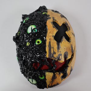 Op de afbeelding: Een tweekleurig Halloween-masker. De ene kant is zwart met meerdere groene ogen, de andere is geel met een zwart kruis en een rode mesh mond. Het masker heeft een getextureerd, grotesk uiterlijk.