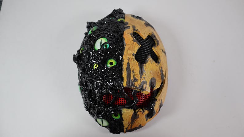 Op de afbeelding: Een tweekleurig Halloween-masker. De ene kant is zwart met meerdere groene ogen, de andere is geel met een zwart kruis en een rode mesh mond. Het masker heeft een getextureerd, grotesk uiterlijk.