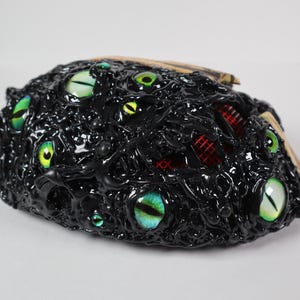 Op de afbeelding: Een zwarte, getextureerde sculptuur met meerdere, kleurrijke, gloeiende ogen. De sculptuur heeft een ruwe, organische uitstraling, met een mix van groene, gele en blauwe ogen. Rode accenten zijn zichtbaar.