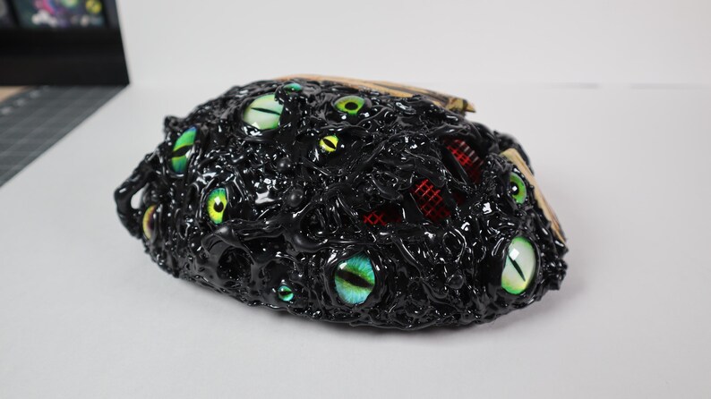 Op de afbeelding: Een zwarte, getextureerde sculptuur met meerdere, kleurrijke, gloeiende ogen. De sculptuur heeft een ruwe, organische uitstraling, met een mix van groene, gele en blauwe ogen. Rode accenten zijn zichtbaar.