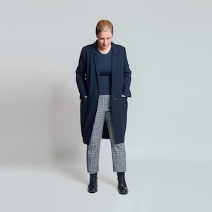 May include: A woman wearing a dark blue long coat with a black shirt and grey and black checked trousers. She is standing with her hands in her pockets and looking down.
