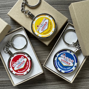 May include: Three keychains with a Vegas-themed design. Each keychain features a round, silver-coloured metal charm with a colourful design. The charms have the text "The Usher" and the names "Spencer", "Kevin", and "Jacob" in a red, yellow, and blue design, respectively. Each charm also includes the date "September 21st 2024" and the names "Megan & Alec".