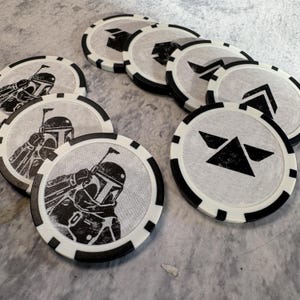May include: A collection of white and black poker chips. Some chips feature a black illustration of a helmeted figure, while others display geometric shapes. The chips have a black and white striped edge.