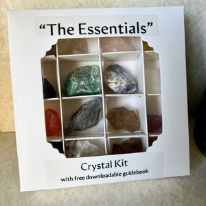 May include: A white square box labeled "The Essentials" and "Crystal Kit with free downloadable guidebook" containing a grid of twelve compartments. Each section holds a raw, unpolished mineral stone in various colors including green, blue, red, and brown.