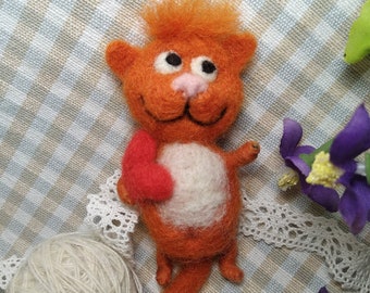 Può includere: Figurina di gatto in feltro arancione fatta a mano con un cuore rosso. Il gatto ha la pancia bianca, occhi neri e naso rosa. Un gomitolo di filo bianco e fiori viola sono sullo sfondo. Il gatto è su un tessuto a scacchi.
