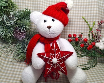 May include: White plush teddy bear wearing a red knit hat and scarf, holding a red star ornament. The bear is sitting on a checkered fabric background.