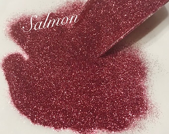 May include: A close-up of a scoop of fine, sparkly, salmon-colored glitter. The glitter is loose and flowing out of the scoop onto a white surface. The word "Salmon" is written in white text on the surface.