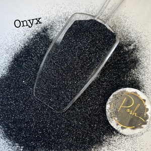 May include: A scoop of black glitter with the word "Onyx" written above it. The glitter is spread out on a white surface and a small round sticker with the word "Posh" is in the lower right corner.