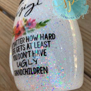 Puede incluir: Vaso blanco brillante con la palabra "Gigi" en escritura negra, detalles florales y el texto "No importa lo dif&iacute;cil que se ponga la vida, al menos no tienes nietos feos". Se ve un logotipo de "Sippin Pretty Designs LLC".